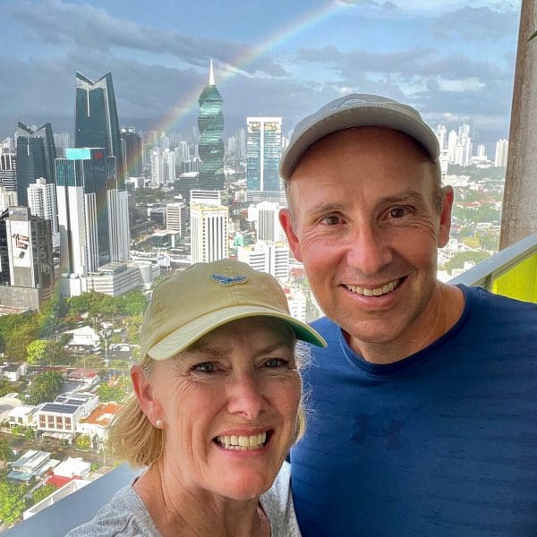 John & Bev, retirement travelers, standing on balcony with Panama City skyline in rear with rainbow