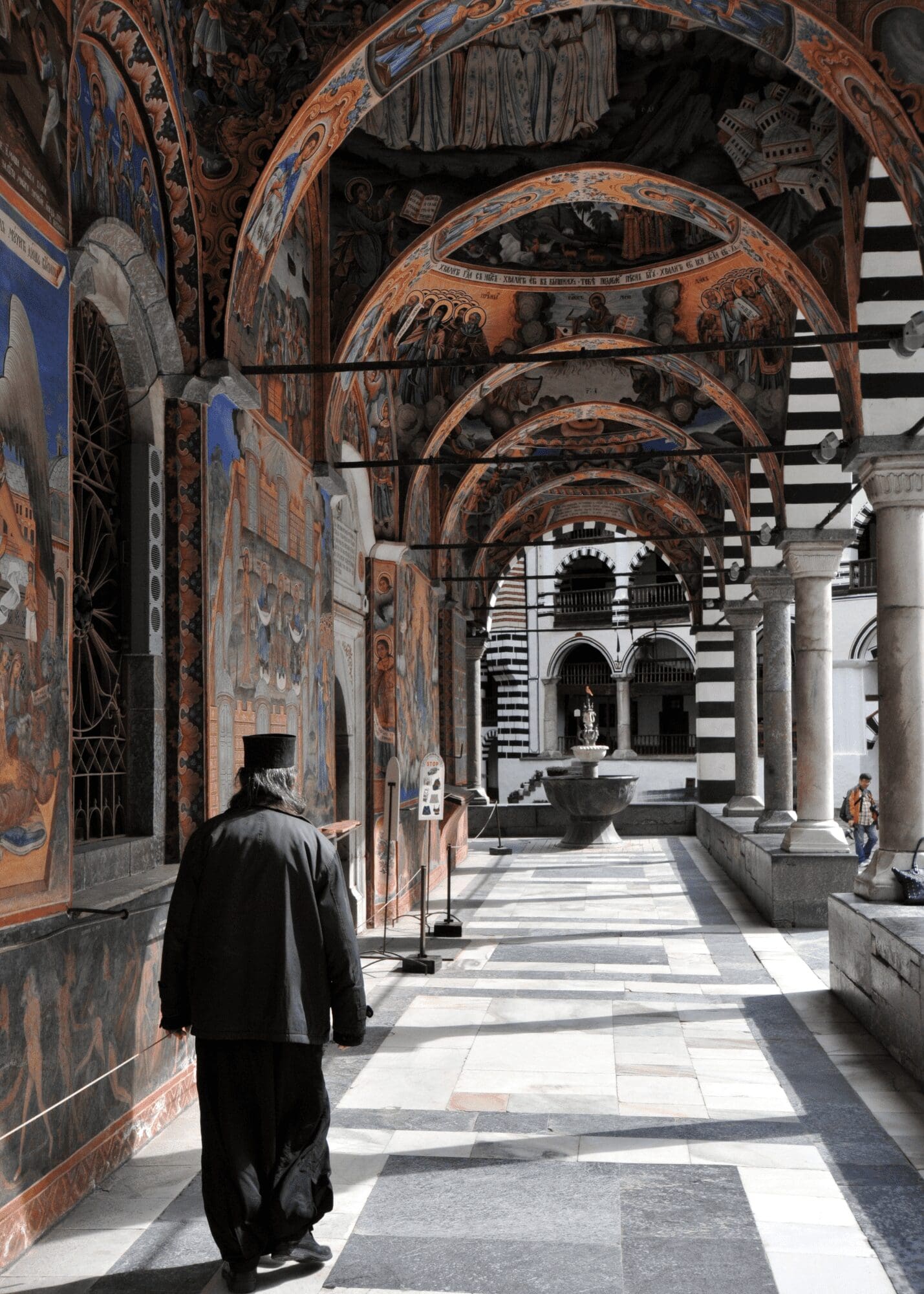 a Priest walking in black garb under an archway of painted columns