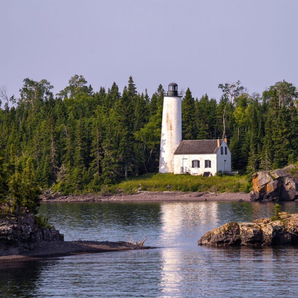 a white lighthouse with white building next to it set along a lake with green pine trees