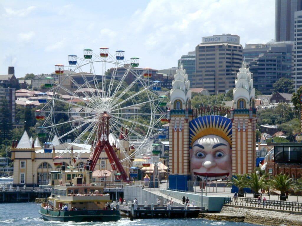 small carnival area with ferris wheel  along a river with a boat passing by