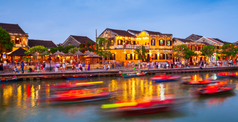 Boats passing through a canal in the city of Hoi An at dusk with restaurants, stores, and people walking next to it.
