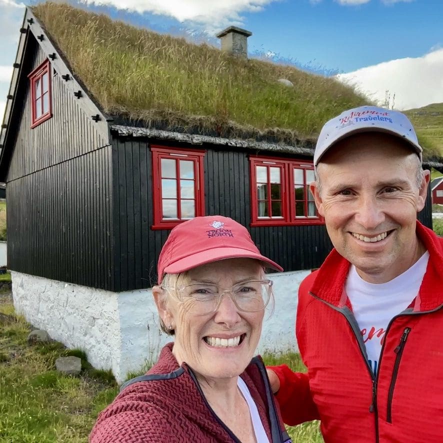 john and bev of the Retirement travelers standing by a turf house in Faroe Islands